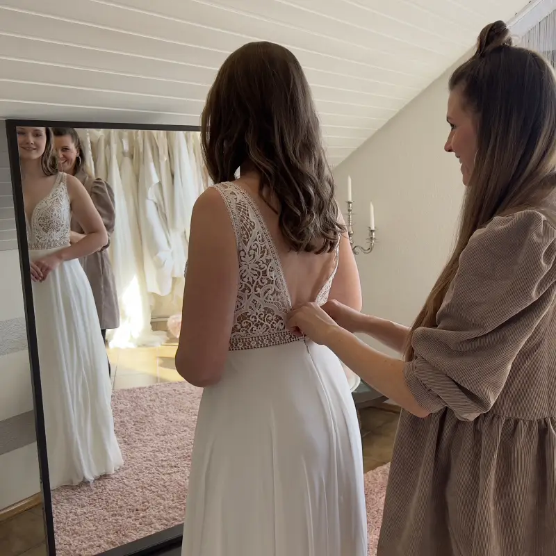 Andrea schließt vor dem Spiegel das Kleid einer Braut