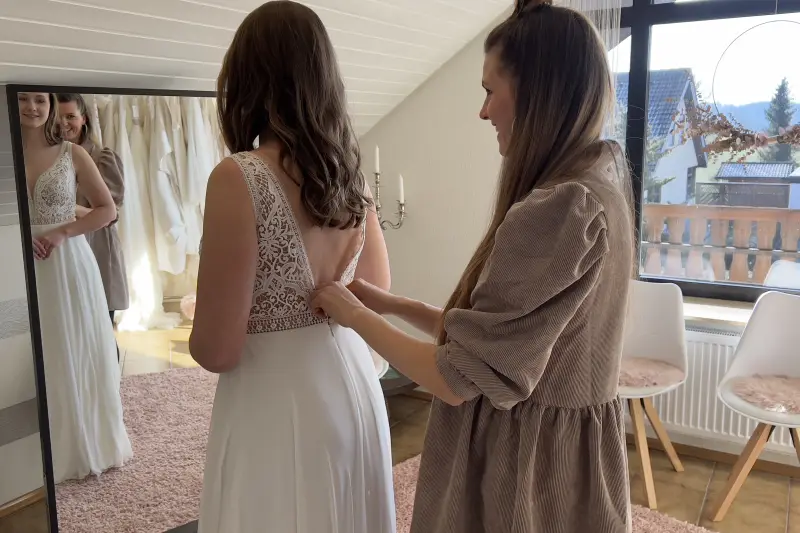 Andrea schließt vor dem Spiegel das Kleid einer Braut
